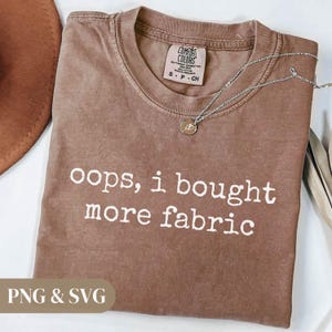 Könnte beinhalten: Ein braunes T-Shirt mit dem weißen Text "oops, i bought more fabric". Das Shirt hat einen Rundhalsausschnitt und ein kleines Etikett oben. Eine silberne Halskette mit Anhänger liegt auf dem Shirt.