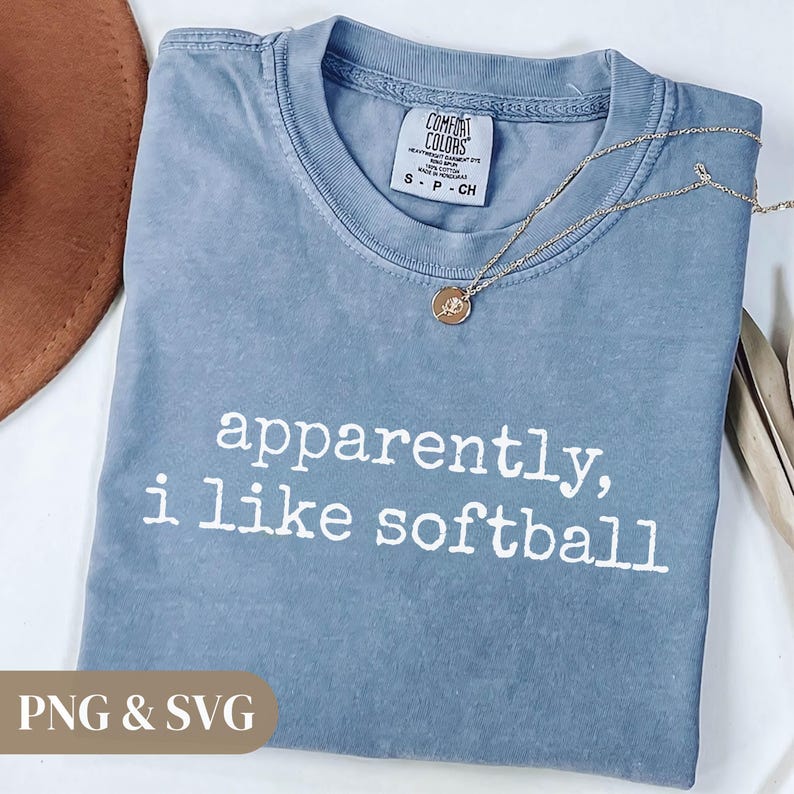 K&ouml;nnte beinhalten: Ein blaues Comfort Colors T-Shirt mit dem wei&szlig;en Text "apparently, i like softball". Eine goldene Halskette mit Anh&auml;nger liegt &uuml;ber dem Shirt. Das Shirt hat ein kleines Etikett am Ausschnitt.