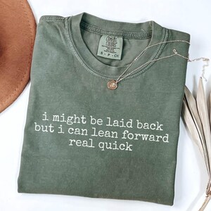 Puede incluir: Camiseta verde oliva con el texto blanco « i might be laid back but i can lean forward real quick ». También se ven un collar con colgante dorado y un sombrero marrón. La camiseta es de la marca Comfort Colors.