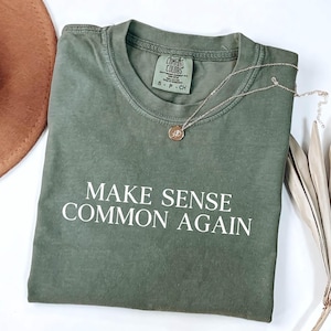 以下が含まれることがあります： 「MAKE SENSE COMMON AGAIN」の白文字が入ったオリーブグリーンのTシャツ。ゴールドのペンダントネックレスと茶色の帽子も見える。シャツは柔らかい素材でできており、サイズはSです。