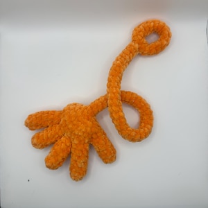 May include: An orange plush hand toy with five fingers and a long, curved arm ending in a loop. The toy is made of a soft, textured material and is set against a white background.