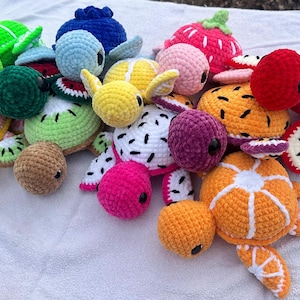May include: A collection of colorful, crocheted fruit-themed turtle plushies. The turtles are in various colors and designs, including orange, green, yellow, pink, and blue, with details like strawberry tops and kiwi slices. The plushies are on a white surface.