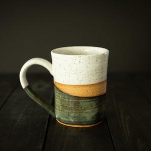 May include: A handmade ceramic mug with a white speckled top, a tan middle section, and a green bottom. The mug has a large, curved handle and is set against a dark background. This stoneware mug is perfect for coffee or tea.