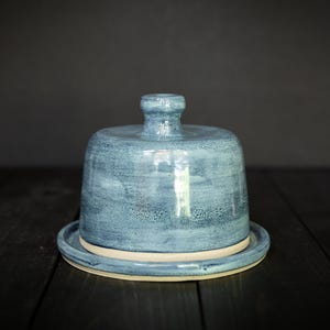May include: A handmade, blue ceramic butter dish with a lid. The dish has a round base and a domed lid with a small handle. The glaze has a mottled appearance, with shades of blue and white. The dish is sitting on a dark wooden surface.