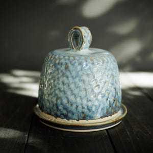 May include: A ceramic butter dish with a dome-shaped lid featuring a textured, dimpled surface in shades of light blue and white. The lid has a small, arched handle at the top, and the base is a flat, circular plate with a smooth, light-colored rim.