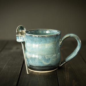 May include: A handmade ceramic mug with a blue glaze and a built-in spoon rest. The mug has a curved handle and a silver spoon. The mug is approximately 4 inches tall.