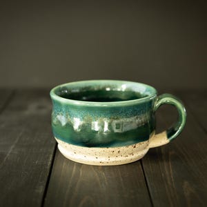 May include: Handmade ceramic mug with a green and white glaze. The mug features a rounded body, a sturdy handle, and a speckled, unglazed base. The interior is dark, suggesting it's ready for coffee or tea.
