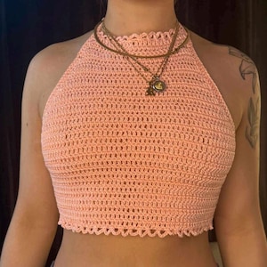 May include: A peach-colored, handmade crochet halter top with a scalloped neckline and hem. The top is worn with a layered gold necklace. The open weave design is visible throughout the garment, showcasing the intricate stitch work.