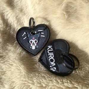 May include: Two heart-shaped keychains with black trim and a black metal ring. One keychain features a cartoon character with Japanese text. The other keychain has the word "KUROMI" in white text. The keychains are on a fluffy, cream-colored surface.