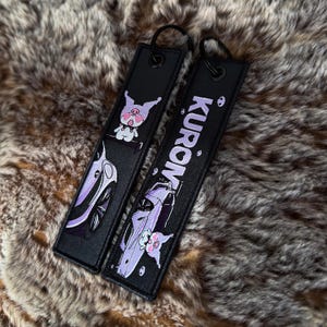May include: Two black keychains with a cartoon pig and car design. One keychain has the word "KUROM" in white. The other keychain has a cartoon pig and car design. Both keychains have a black loop.