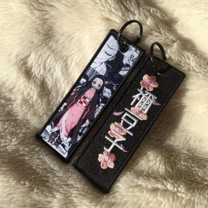 May include: Two rectangular keychains. One features a character in a pink kimono and black jacket. The other is black with embroidered Japanese characters and pink flowers. Both have black metal rings.