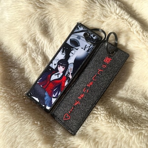 May include: Two rectangular keychains. One features an anime character in a red jacket, white shirt, and blue skirt. The other is black with red Japanese text and a heart. Both have metal key rings.