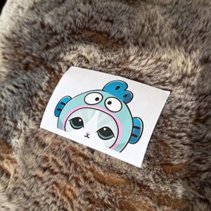 May include: A white sticker featuring a cartoon cat wearing a blue fish costume. The cat has large, expressive eyes and a small pink nose. The sticker is on a textured, brown surface.