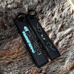May include: Two black keyrings with metal hoops. One has a teal and black anime-style illustration with the word "DARLINGFUL" in teal. The other has a teal and white abstract design. Both are on a brown, furry surface.