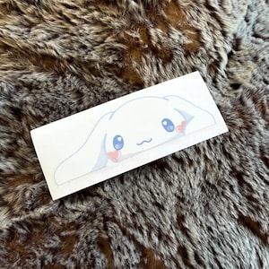 May include: A rectangular white sticker featuring a cartoon character with long white ears, large blue eyes, a small mouth, and pink heart-shaped cheeks. The character is depicted in a peeking pose, with the ears extending horizontally across the sticker.