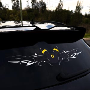 May include: A black and yellow Umbreon character decal with white tribal-style accents applied to the rear window of a black vehicle. The design features the character's head with yellow ear markings and a yellow circular forehead detail centered.