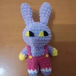 Puede incluir: Un juguete de peluche de conejo tejido a ganchillo con hilo morado claro, con orejas largas, manos amarillas y un overol rosa con dos pequeños botones amarillos. El conejo tiene ojos grandes, planos, amarillos y negros y detalles blancos.