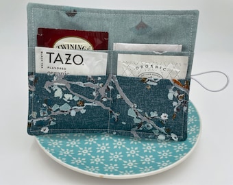 May include: A fabric tea bag organizer with a light blue top section and a dark teal bottom section featuring a white and grey floral branch pattern. The organizer has two front pockets containing Tazo and Archer Farms tea bags.