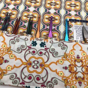 May include: A white fabric roll-up case with a colorful geometric pattern. The case is open and filled with knitting needles and crochet hooks. There is also a pair of scissors and a ruler inside the case.