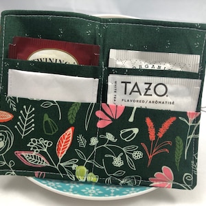 May include: A green fabric tea bag holder with a floral pattern. The holder has four pockets for tea bags. The holder is sitting on a blue and white plate.