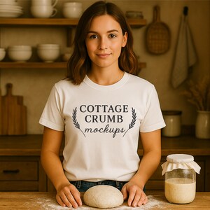 May include: A woman wearing a white t-shirt with the text "COTTAGE CRUMB mockups" in black script and a laurel wreath design. She is standing in a kitchen with a ball of dough and a jar of starter on a wooden surface.