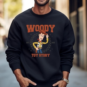 May include: Black sweatshirt featuring a graphic of Woody from Toy Story. The design includes Woody's image, the word "WOODY" in orange, and "TOY STORY" in white. The sweatshirt also has the text "EST. 1995".