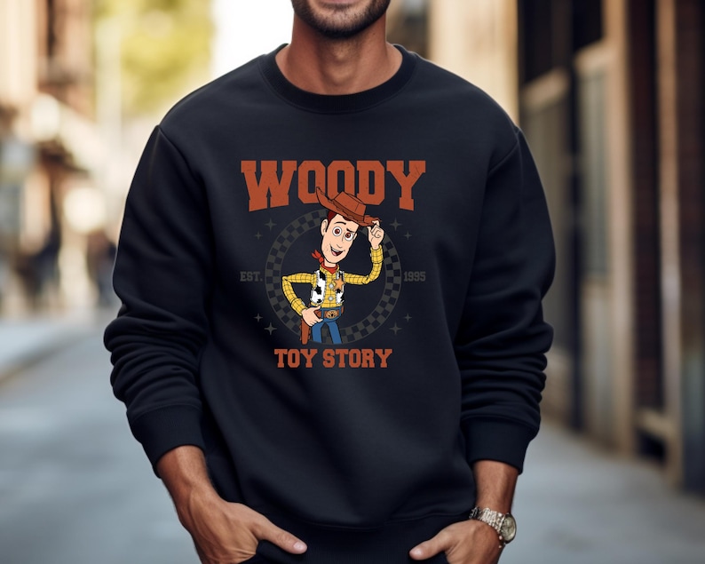 May include: Black sweatshirt featuring a graphic of Woody from Toy Story. The design includes Woody's image, the word "WOODY" in orange, and "TOY STORY" in white. The sweatshirt also has the text "EST. 1995".