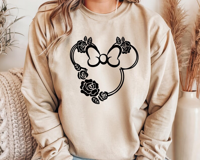 May include: Beige sweatshirt featuring a black floral Mickey Mouse design. The design includes a bow and roses. The sweatshirt is a neutral color and has long sleeves. The design is centered on the front of the shirt.
