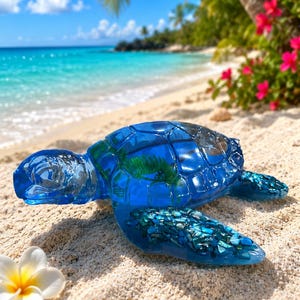 May include: A translucent blue sea turtle figurine crafted from resin, featuring a detailed shell filled with green foliage and small blue stone fragments embedded within the flippers, resting on a sandy beach near a white plumeria flower.