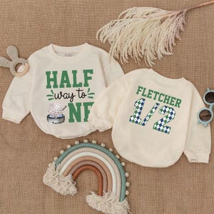 May include: Two cream-colored baby sweatshirts. One has the text "HALF way to ONE" in green, with a golf ball graphic. The other says "FLETCHER 1/2" in green and blue checkered pattern. A rainbow and sunglasses are also in the image.