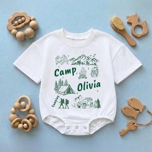 May include: White baby onesie with a green camping-themed graphic. The design includes mountains, trees, a tent, a campfire, and a jeep. The text "Camp Olivia" is printed in green. Wooden toys and a brush surround the onesie.