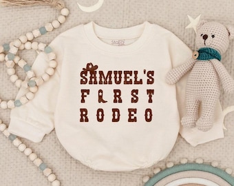 May include: Cream-colored baby onesie with the text "SAMUEL'S FIRST RODEO" in brown. A cowboy hat and boot graphic are above the text. A small stuffed bear with a teal scarf is next to the onesie.