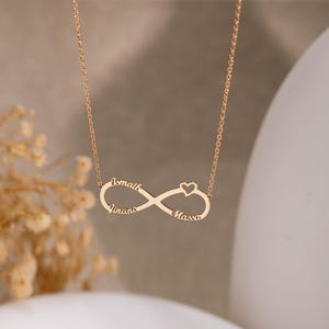 Op de afbeelding: Een goudkleurige ketting met een oneindigheidssymbool en de namen Ismaik, Jinans en Massa. Een klein hartje is rechts van het oneindigheidssymbool geplaatst. De ketting hangt aan een fijne ketting.
