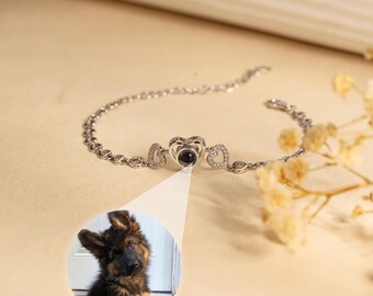 May include: A silver-colored bracelet with a heart-shaped pendant. The pendant has a small, dark circle in the center. Two smaller heart-shaped charms flank the pendant. A close-up of a German Shepherd puppy is in the lower left corner.