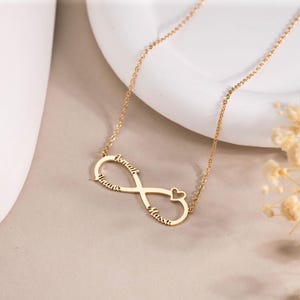 May include: A gold-colored infinity necklace with the names "Imane", "Ismail", and "Massa" engraved on it. The necklace features a small heart design and is displayed on a white surface.