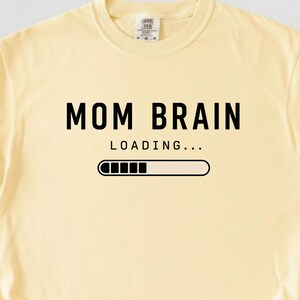 May include: A pale yellow t-shirt featuring the words "MOM BRAIN" in black, above "LOADING..." and a loading bar graphic. The shirt is made of a soft material and has short sleeves.