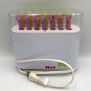 May include: A white Conair Hot Sticks hair styling appliance with a clear plastic lid. The unit features a row of alternating light green and purple plastic styling wands inserted into the base. A white power cord with a plug is coiled in front.