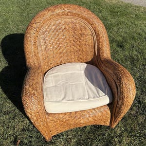May include: A rounded armchair constructed from woven light brown wicker with a herringbone pattern. The chair features a high, curved backrest, integrated armrests, and a removable, flat, off-white fabric seat cushion centred on the base.
