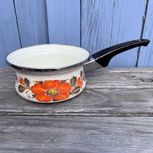 May include: A vintage enamel saucepan with a black handle and a floral design. The pot features a cream-colored interior and a speckled exterior with orange and brown flowers. The handle is black and the rim is silver.