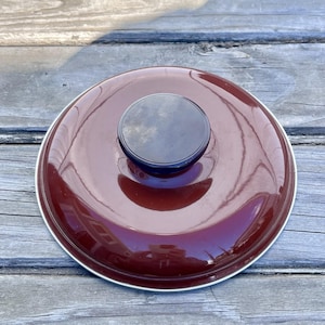 May include: A round, maroon-colored pot lid with a black, circular handle. The lid has a shiny, reflective surface and a silver-colored rim. The lid is sitting on a weathered, wooden surface.