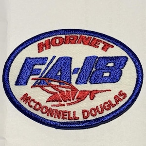May include: An oval-shaped embroidered patch with a white background and a blue border. It features the red text HORNET at the top, large blue text F/A-18 in the center, a red hornet graphic, and the red text MCDONNELL DOUGLAS at the bottom.