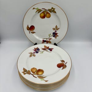 Puede incluir: Una pila de platos de cerámica blanca con borde dorado. Cada plato presenta un diseño pintado de frutas, incluyendo peras, melocotones y moras. Los platos están dispuestos sobre un soporte negro sobre un fondo blanco.