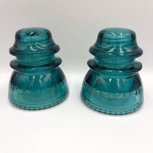 May include: Two teal-colored glass insulators with a stepped, dome-shaped top and a wide, flared base. Each piece features embossed text reading HEMINGRAY-42 along the lower edge, with a textured, beaded pattern circling the bottom rim.