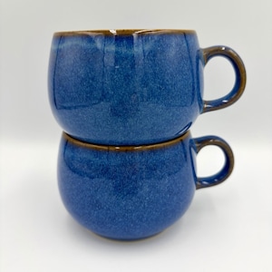 Puede incluir: Dos tazas redondas de cerámica azul apiladas con bordes y asas marrones. Las tazas tienen una textura moteada y un acabado brillante. Las asas son de color marrón a juego.
