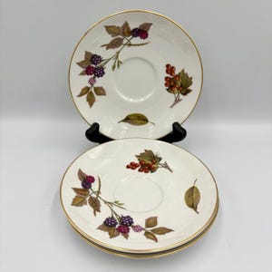 Puede incluir: Dos platillos de cerámica blanca con bordes dorados, con un diseño botánico de moras y hojas en tonos morados, marrones y verdes. Los platillos están apilados sobre un soporte negro.