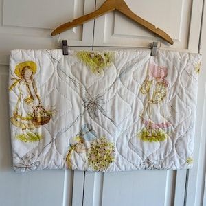 Puede incluir: Tela acolchada blanca con ilustraciones de niños con gorros y vestidos, llevando cestas y flores. La tela presenta un patrón de diamantes con acentos de cinta azul claro y está colgada de una percha de madera.