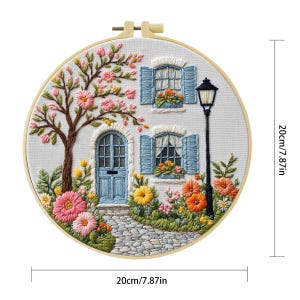 以下が含まれることがあります： 青い窓とドアのある白い家、花と木に囲まれた刺繍フープアート。アートワークは直径20cmで、木製のフープに入っています。