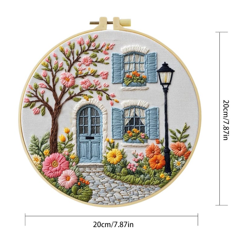 以下が含まれることがあります： 青い窓とドアのある白い家、花と木に囲まれた刺繍フープアート。アートワークは直径20cmで、木製のフープに入っています。