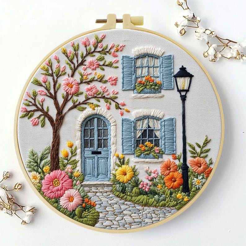 以下が含まれることがあります： 円形に刺繍されたアート作品。青い窓とドア、咲き誇る木、石畳の小道がある可愛らしいコテージのシーンを描いています。カラフルな花と緑でいっぱいのシーンは、鮮やかなディスプレイを作り出しています。