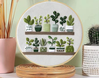 May include: Embroidered hoop art showing a selection of potted plants on shelves. The plants are in various shades of green and are displayed in decorative pots. The embroidery is set against a white fabric background, framed by a wooden hoop.